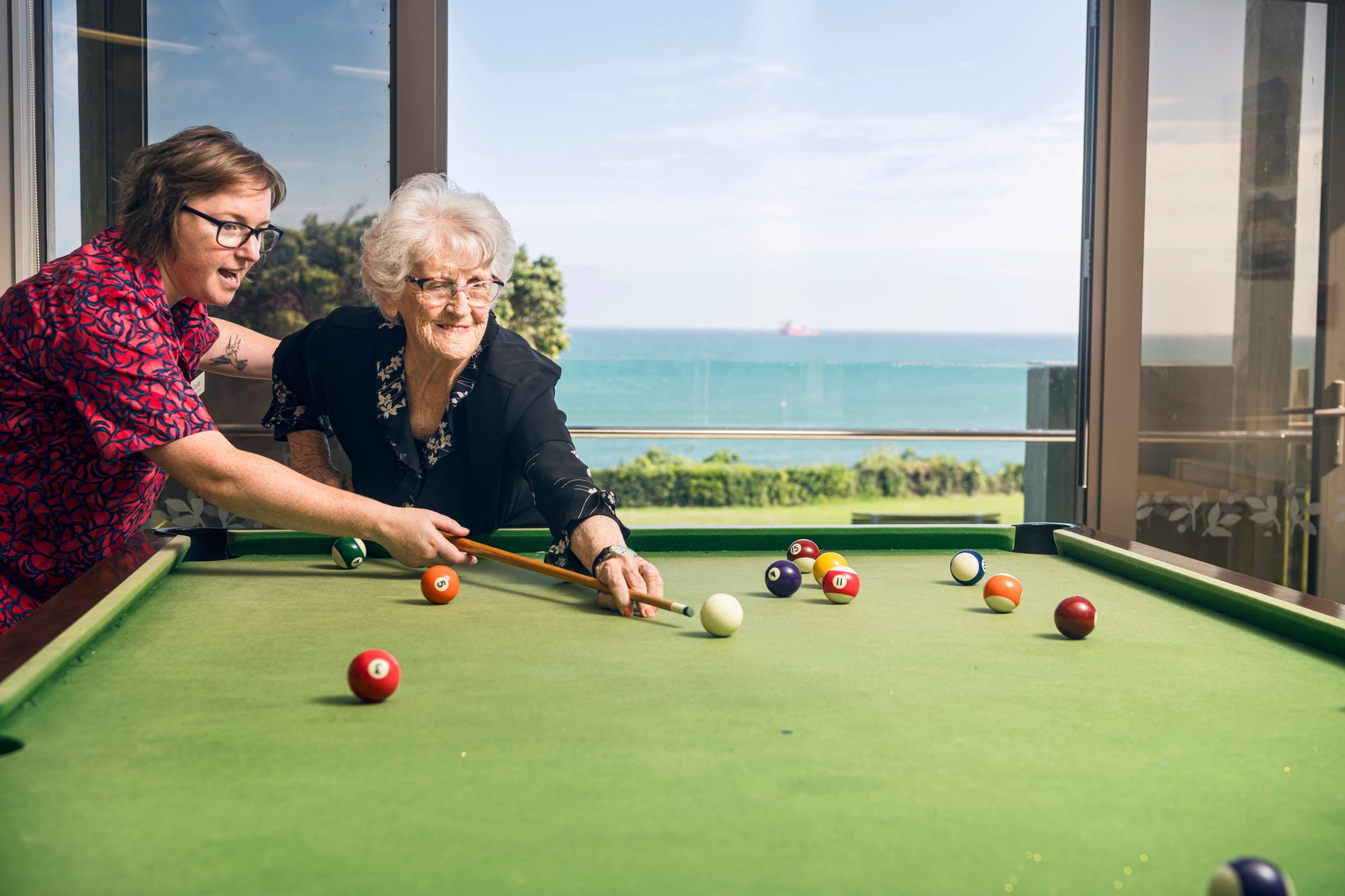 Older woman playing pool in front of ocean with Enliven Staff Member