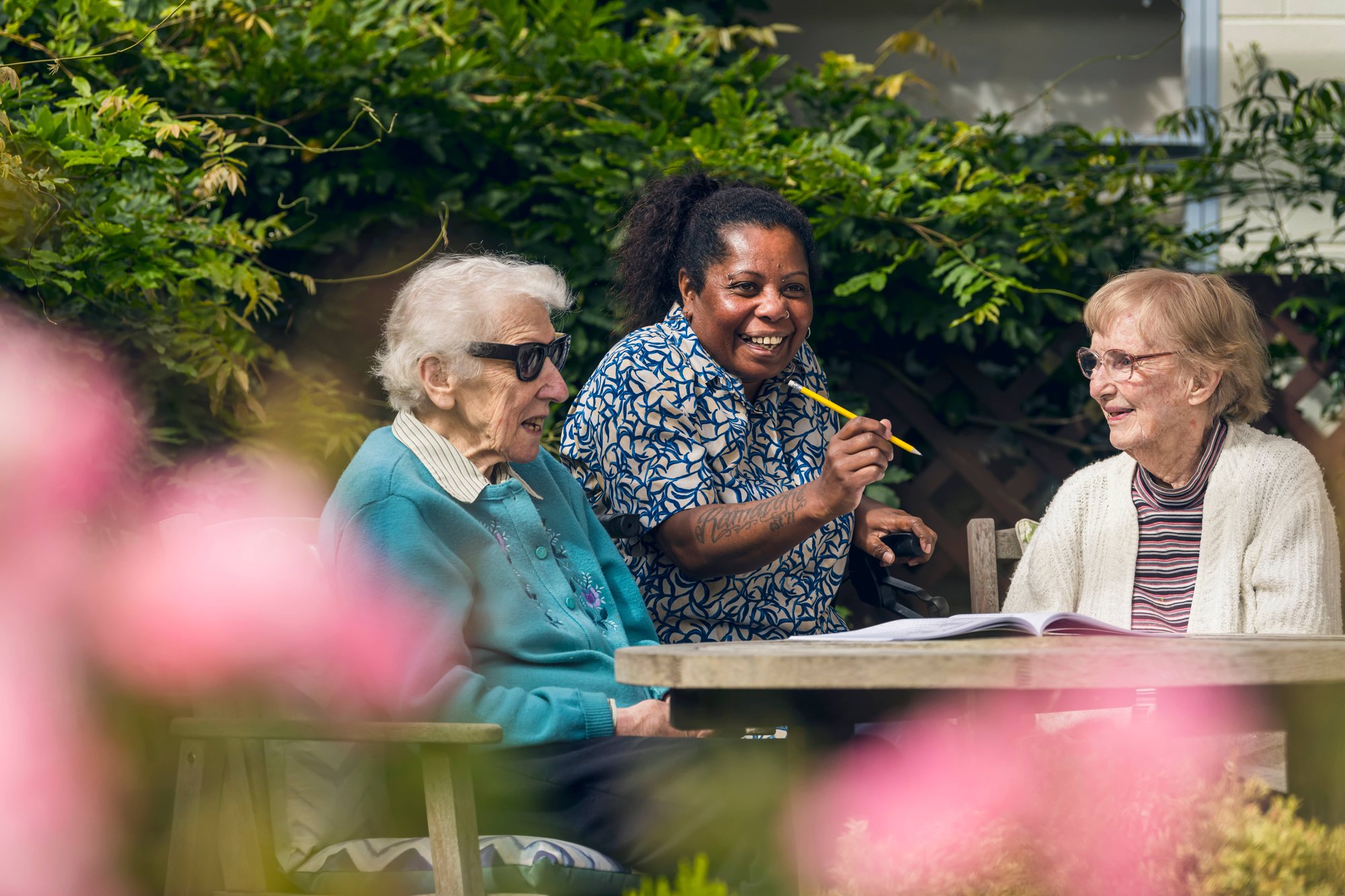 Two older women sitting outside, Enliven staff member holding pen, all smiling